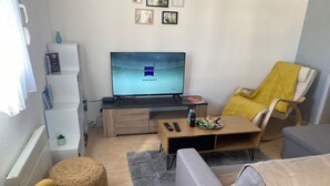 Una sala de estar moderna con un televisor de pantalla plana, una mesa de centro de madera, un sofá, un sillón amarillo y una mesita auxiliar con aperitivos.