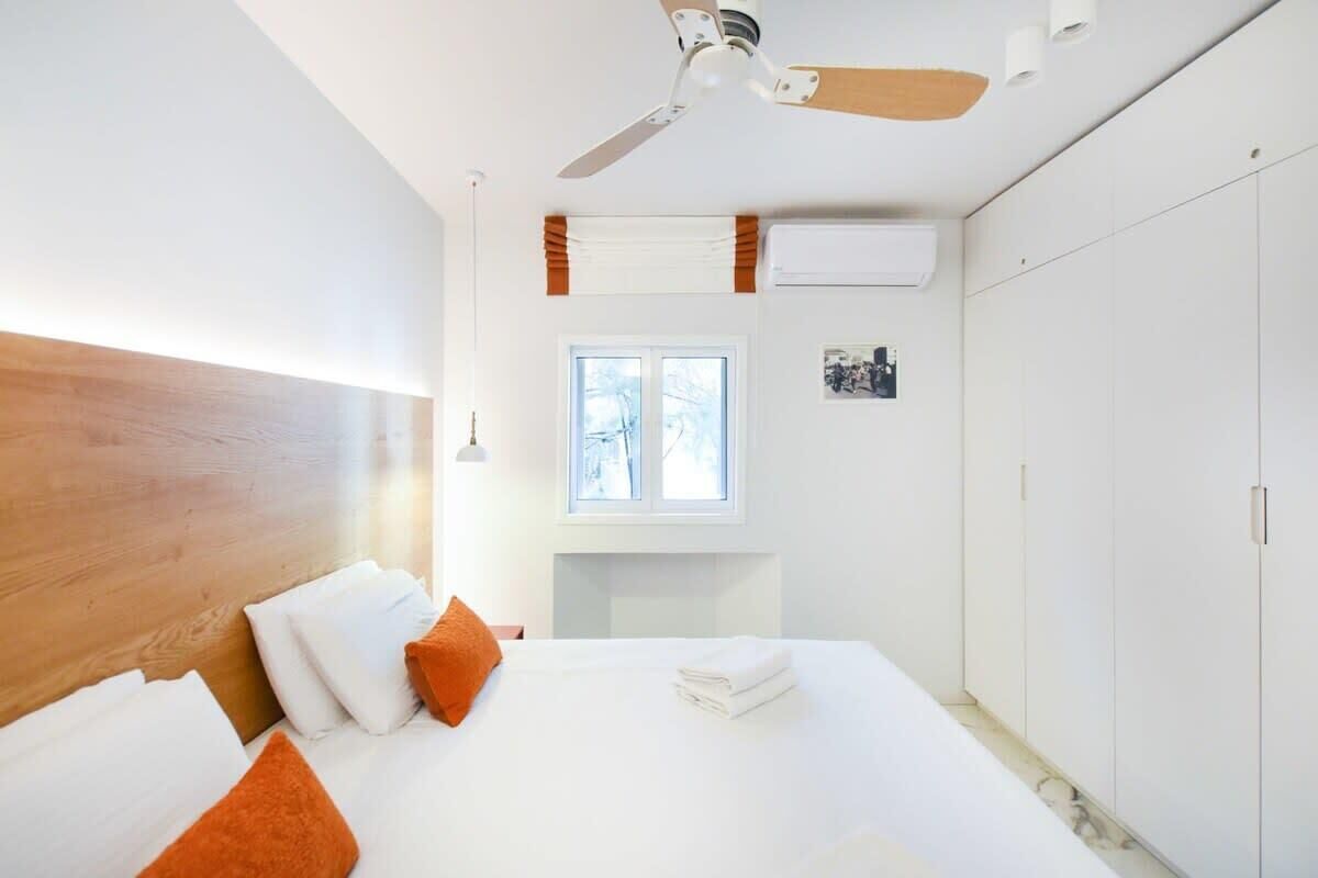 Ett hotellrum med en sänggavel i trä, vita sängkläder, en orange kudde, en takfläkt, ett fönster med utsikt och en vit garderob.