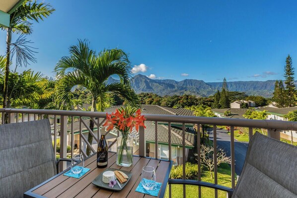 A balcony with a table set for two, featuring a bottle of wine, a glass, a vase with flowers, and a plate with food. The balcony overlooks a lush landscape with mountains in the distance. - Villas Of Kamalii 55: 3 BR, 3 BA Condominium in Princeville, Sleeps 6 (Princeville)