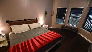 Une chambre à coucher comprenant un lit, des tables de chevet, une commode, un banc et des fenêtres équipées de stores.