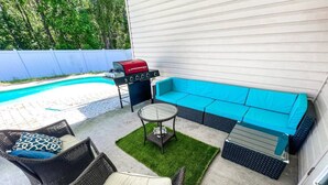 Une terrasse extérieure avec une piscine, un barbecue rouge, un canapé bleu, des fauteuils en osier et une table en verre.