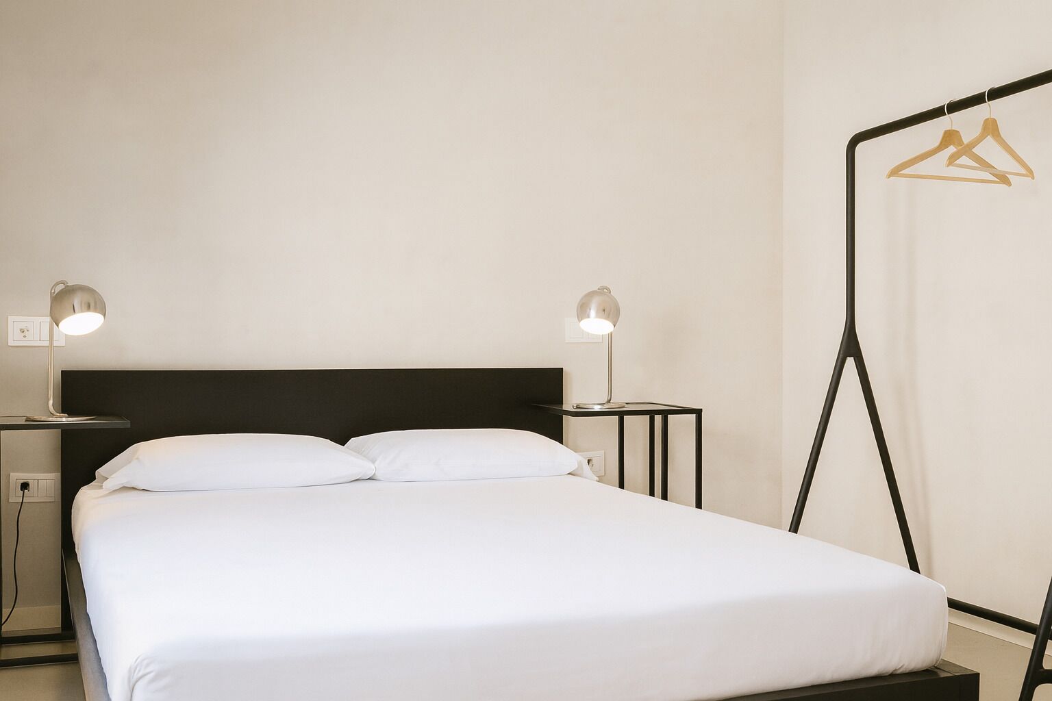 A neatly made bed with white linens, two bedside tables with lamps, and a clothing rack against the wall.