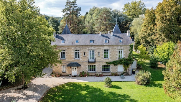 A large stone building with multiple windows and a prominent roofline, surrounded by lush greenery and trees.