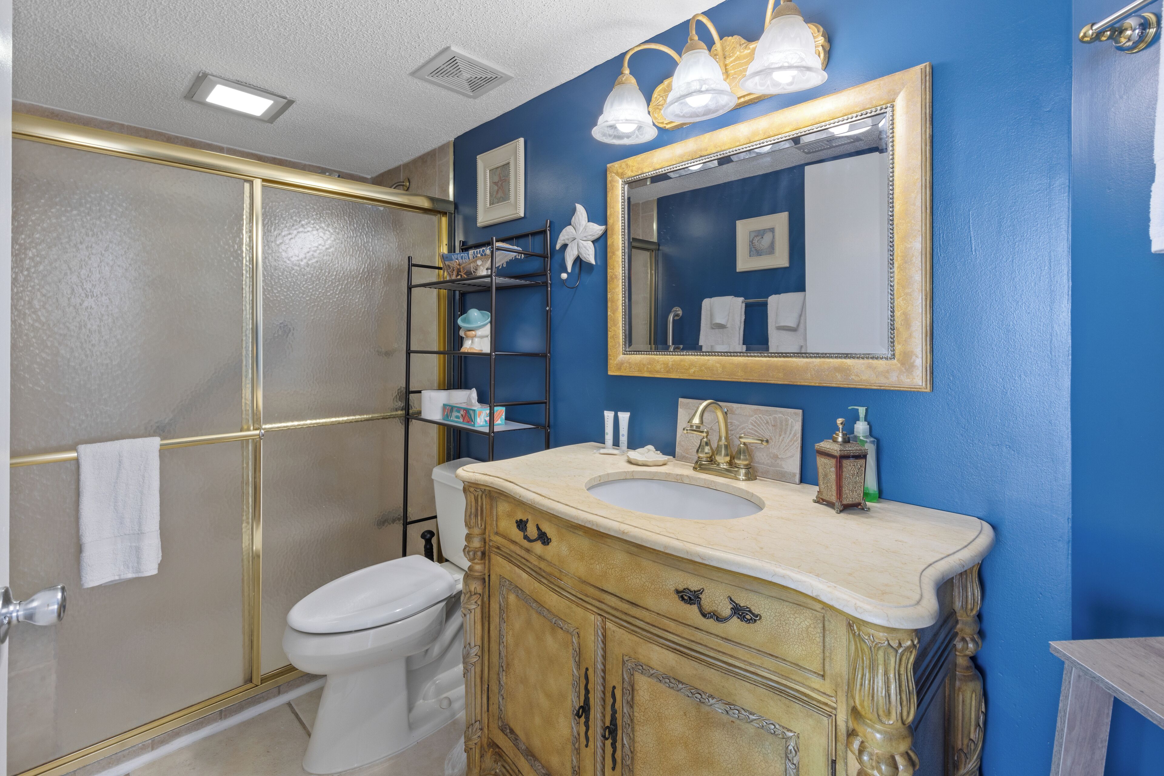 Ein Badezimmer mit einer weißen Toilette, einem beige farbenen Waschbecken mit verziertem Schrank, einer blauen Wand und einem goldfarbenen Spiegel.