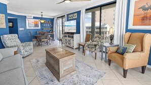 Un vaste salon comprenant une table basse en bois, un canapé et un fauteuil à motifs. On y trouve également un coin repas avec une table et des chaises, ainsi qu’un téléviseur posé sur un meuble. La pièce est dotée de grandes fenêtres avec des rideaux, d’un ventilateur de plafond et de tableaux décorant les murs.