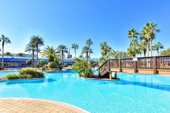 A swimming pool with a wooden deck, palm trees, and a building with blue awnings.