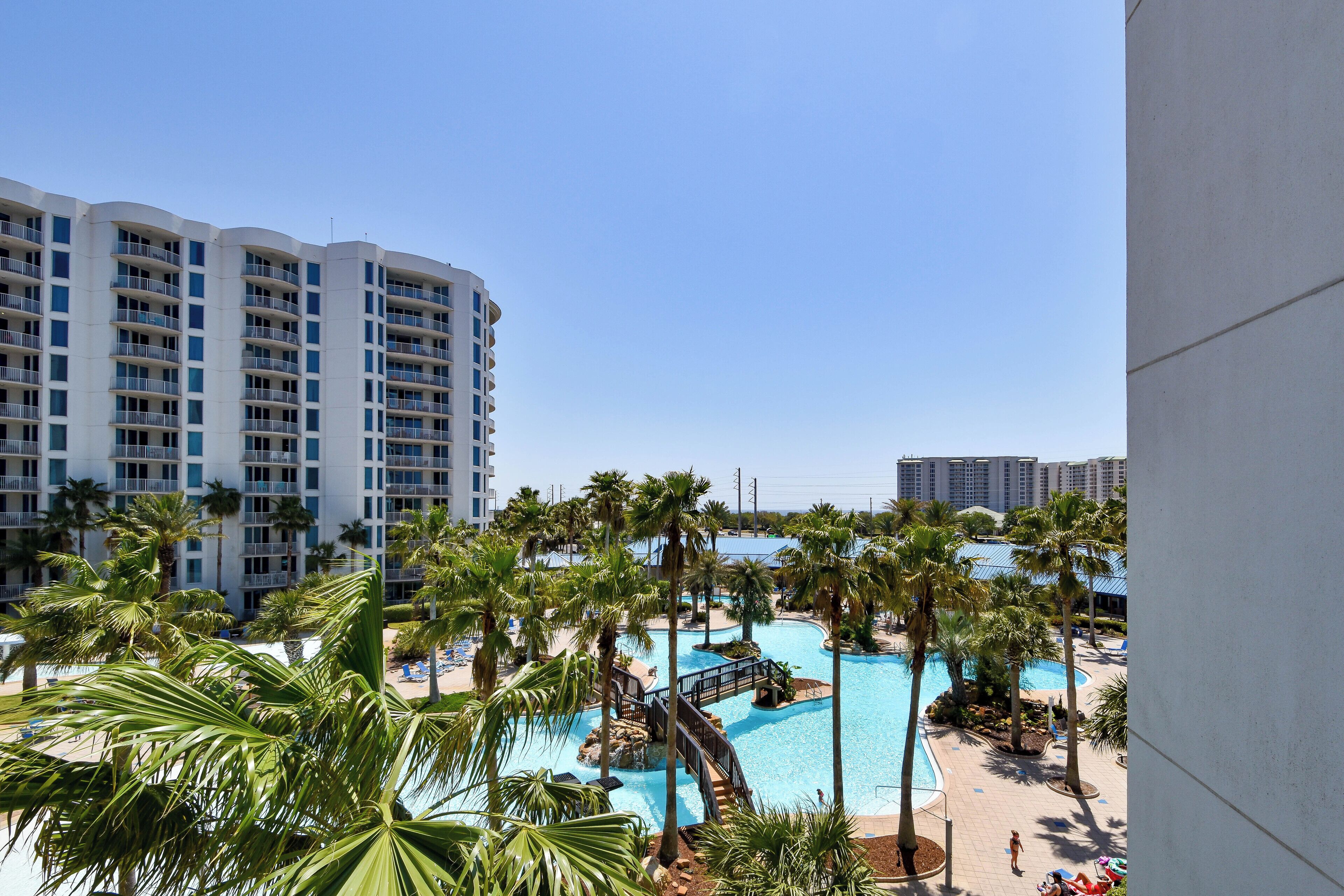 A resort with a swimming pool surrounded by palm trees, a bridge, and a view of high-rise buildings.