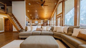 A spacious living room with a large sectional sofa, a wooden staircase, and large windows offering a view of the outdoors. - Flathead River Retreat For 15! (Columbia Falls)