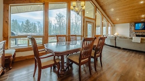 A spacious dining area with a large wooden table and chairs, large windows offering a view of a snowy forest, and a cozy living area with a fireplace. - Flathead River Retreat For 15! (Columbia Falls)