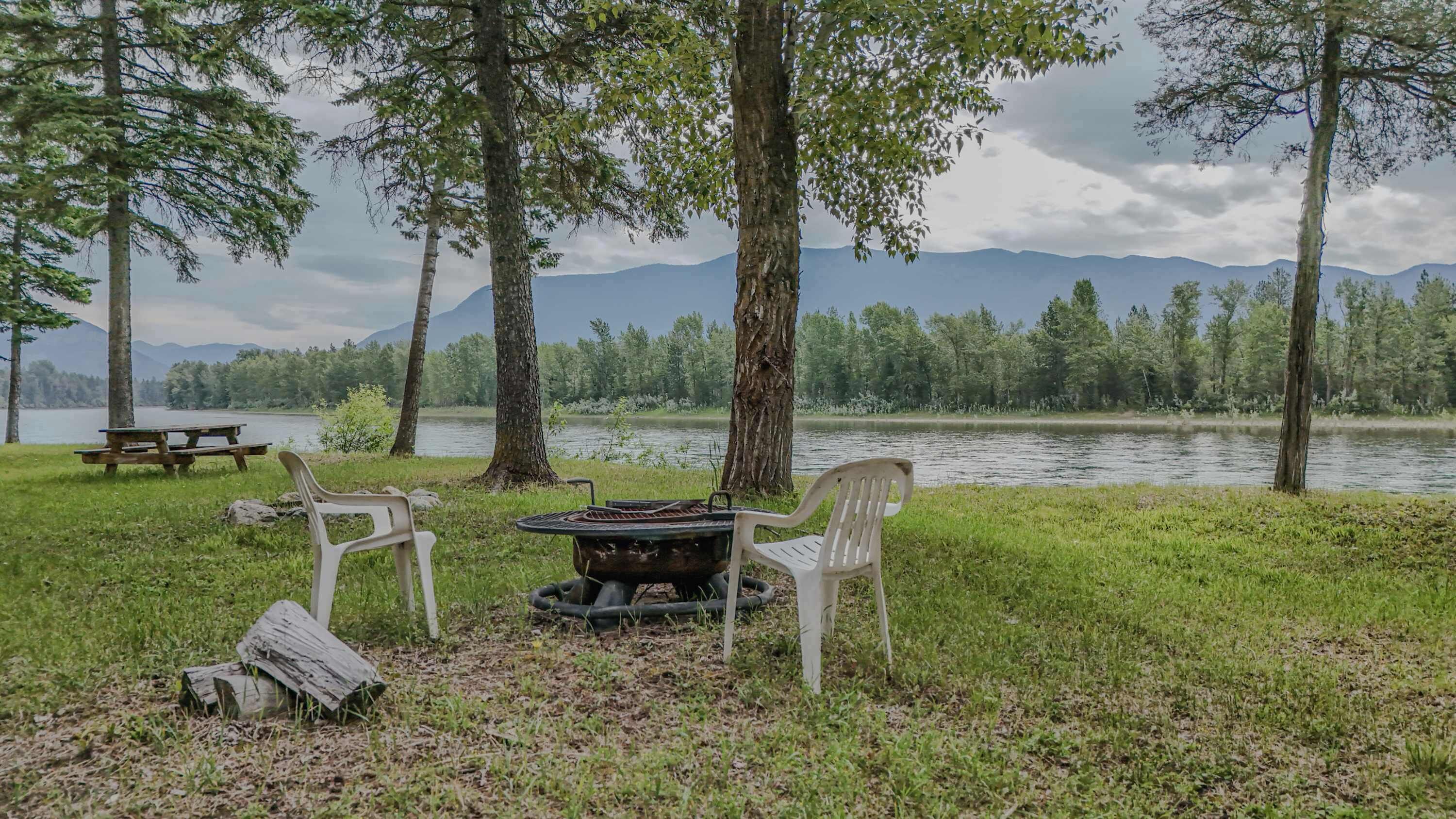 A picnic setup with a grill, chairs, and a table by a lake with mountains in the background.