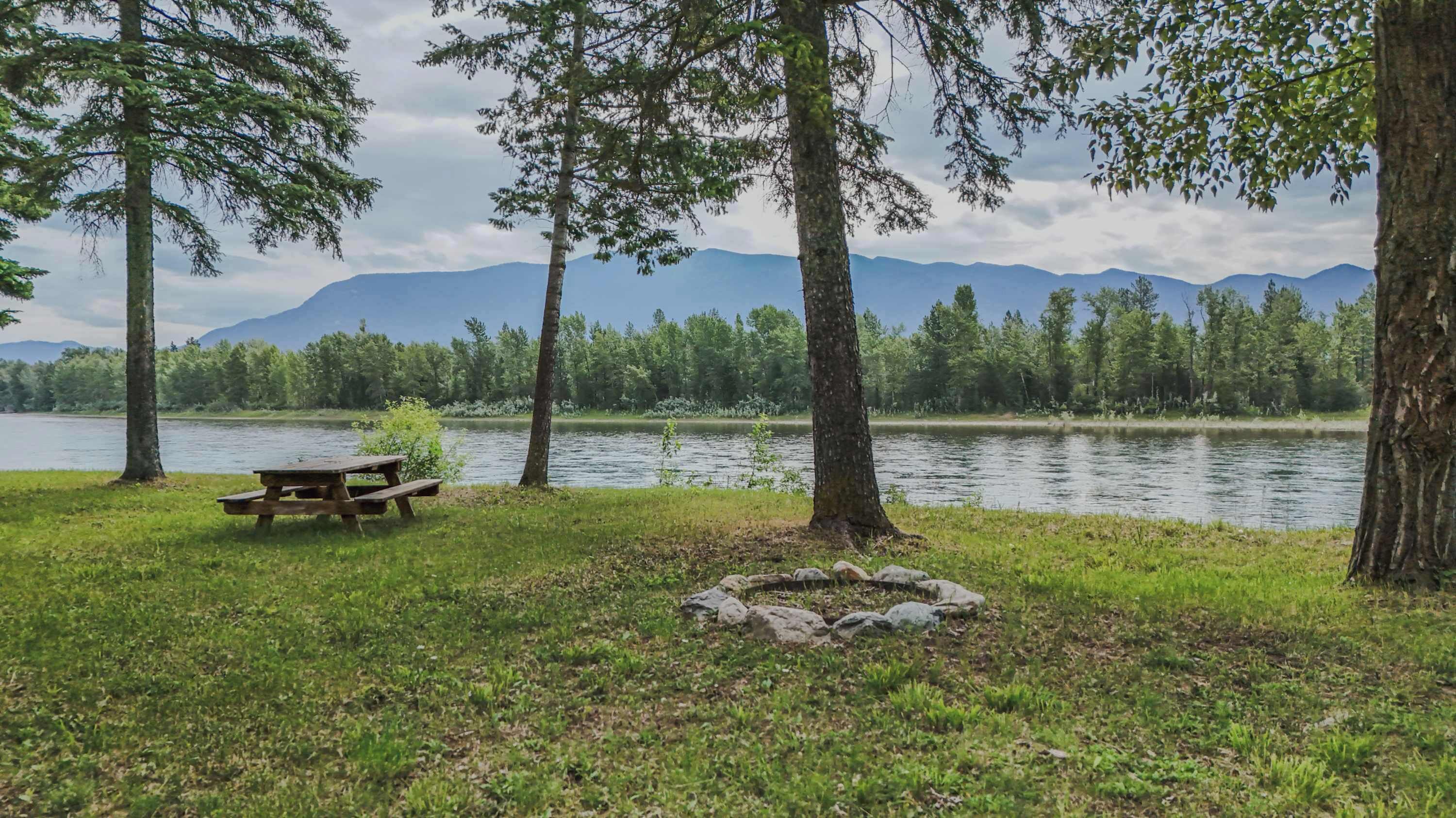 Zona de picnic junto al lago con una mesa y rocas, rodeada de árboles y montañas al fondo.