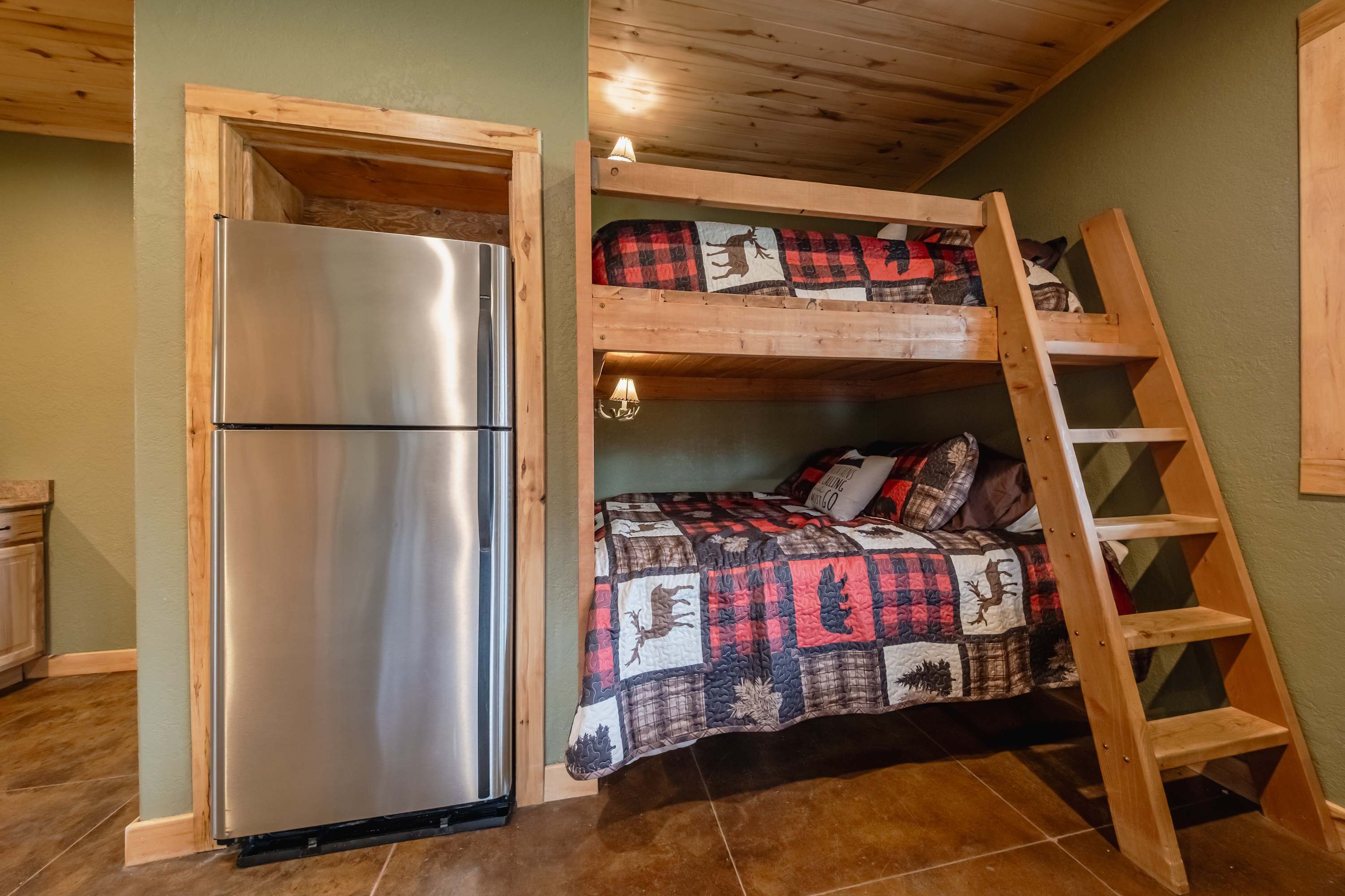 A compact room with a built-in stainless steel refrigerator, a bunk bed with a quilted blanket, and a wooden ladder.
