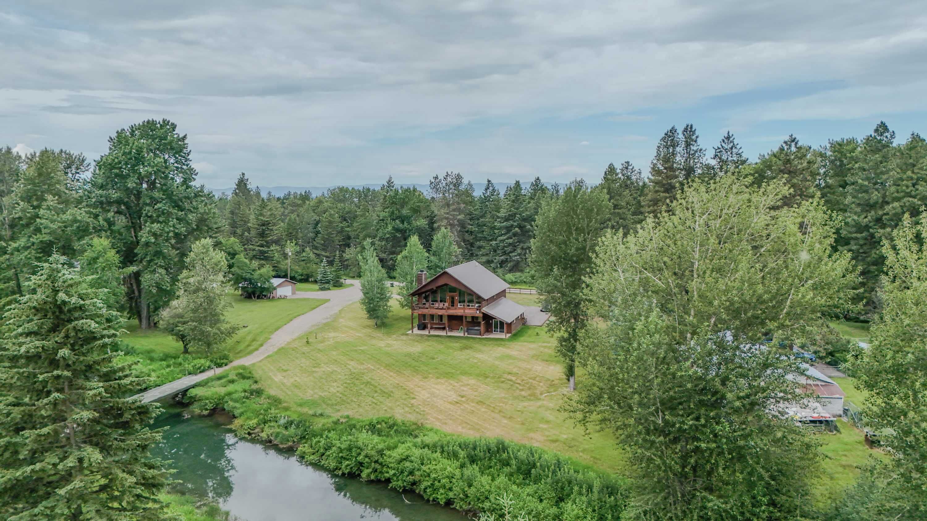 Una cabaña grande de troncos con una terraza, rodeada de un bosque denso y un pequeño arroyo.