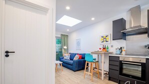 A modern kitchen with a built-in oven, a dining area with a table and chairs, and a blue sofa with colorful cushions. - Host & Stay | Croydon Road Abode (Caterham)