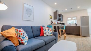 A modern living room with a grey sofa, colorful cushions, a white coffee table, and a kitchen area in the background. - Host & Stay | Croydon Road Abode (Caterham)
