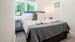 Een netjes opgemaakt bed met witte lakens en een grijze deken, een bedlamp en een lijst met een foto aan de muur.