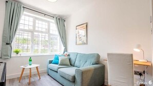 Een gezellige kamer met een lichtblauwe bank, een witte bijzettafeltje, een lamp en een raam met gordijnen.