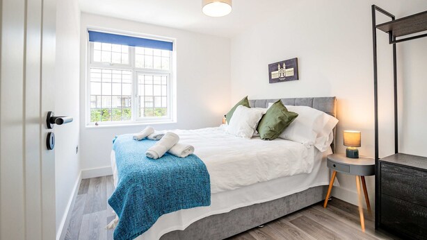 Un dormitorio con cama, una mesita de noche con lámpara, una ventana con persianas y una puerta.