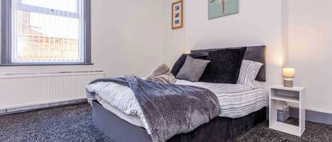 A bedroom with a bed, a bedside table, a window, and a wall with a picture.