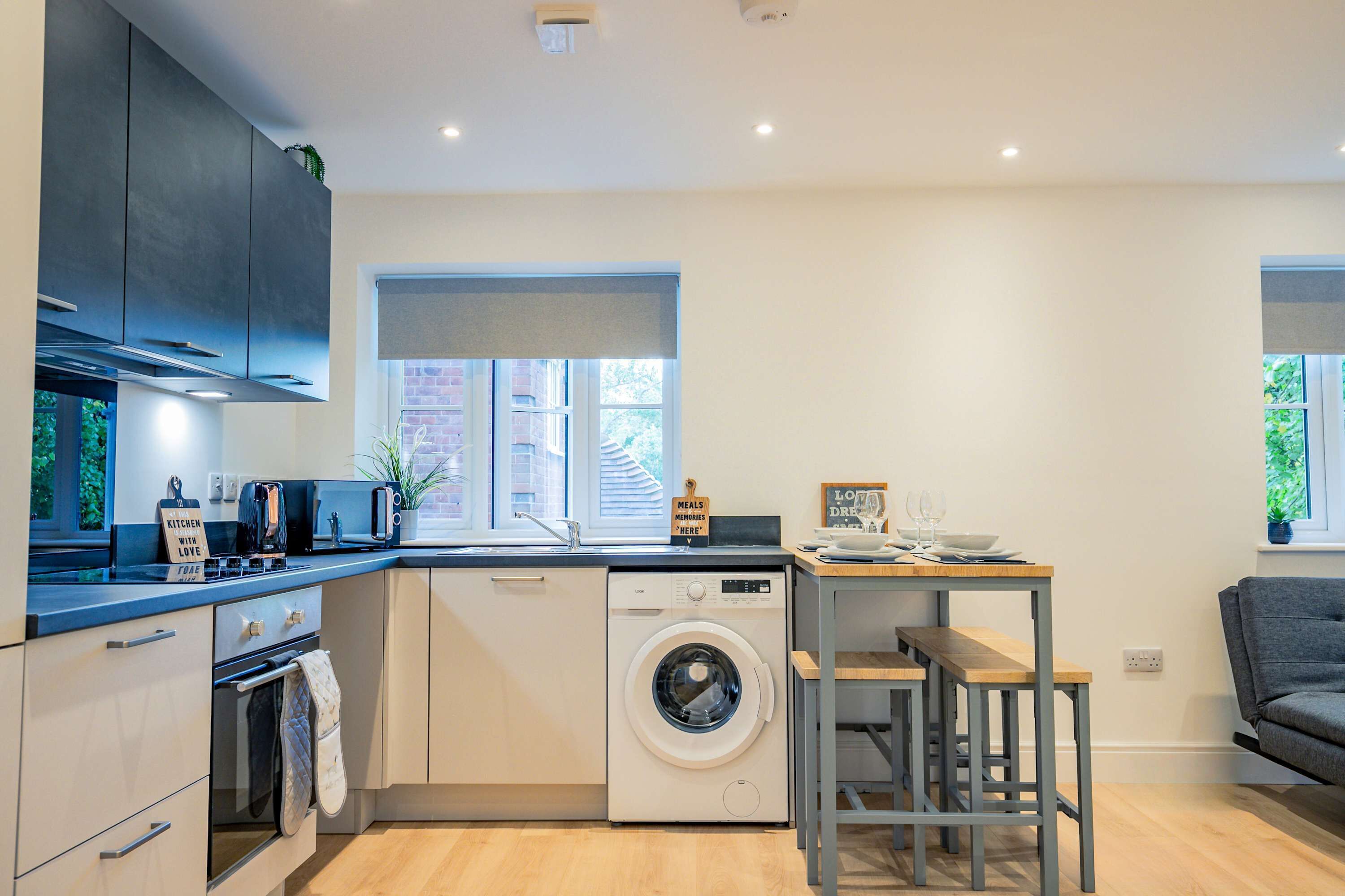 A compact kitchen with a washing machine, a dining area with a table and chairs, and a grey sofa.