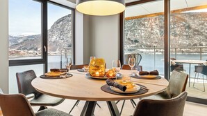 A dining area with a round table, chairs, and a large window offering a view of snow-covered mountains and a body of water. - Penthouse with amazing fjord view (Vestland)