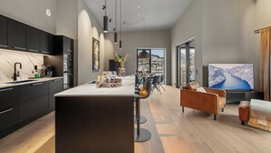 A modern kitchen with dark cabinetry, a central island, and pendant lighting. There is a dining area with a sofa and chairs, and a large window offering a view of a snowy landscape. - Penthouse with amazing fjord view (Vestland)