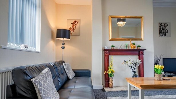 Un salon avec un canapé en cuir noir, une table basse en bois, un lampadaire, une cheminée surmontée d’un miroir et un tableau encadré accroché au mur.