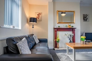 Een woonkamer met een zwarte leren bank, een houten salontafel, een staande lamp, een open haard met een spiegel erboven en een inlijsting foto aan de muur.
