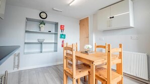 Une cuisine moderne avec une table et des chaises en bois, des meubles blancs, une horloge et une petite étagère ornée d’objets décoratifs.