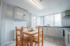 A modern kitchen with white cabinets, a wooden dining table, and a window with blinds. - Host & Stay | Park Cliffe House (Bradford)