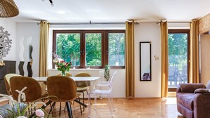 Een ruime woonkamer met een eettafel, stoelen en een bank. Grote ramen bieden uitzicht op groen buiten.