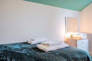 A neatly made bed with a green blanket, a bedside table with rolled towels, and a nightstand with a lamp.
