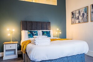 A neatly made bed with white linens and a blue accent throw, a small bedside table with a lamp, and a wall with two framed artworks.