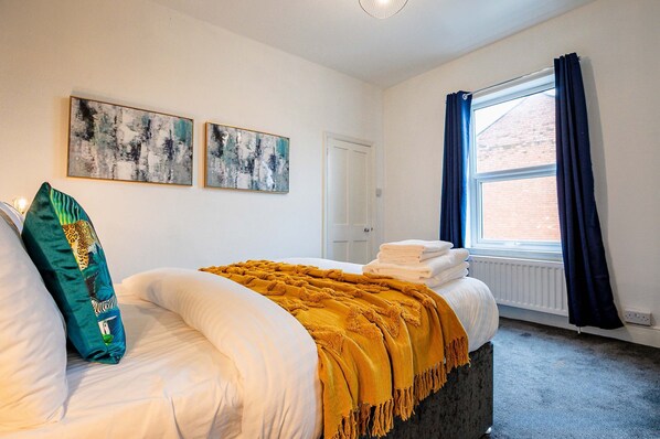 A bedroom with a bed, two wall-mounted abstract paintings, a window with blue curtains, and a radiator.