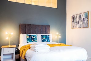 A neatly made bed with white linens and a yellow throw blanket, a bedside table with a lamp, and a wall-mounted abstract painting.