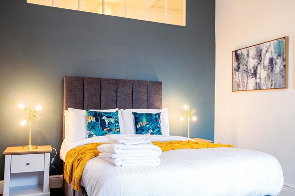 A neatly made bed with white linens and a yellow throw blanket, a bedside table with a lamp, and a wall-mounted abstract painting.