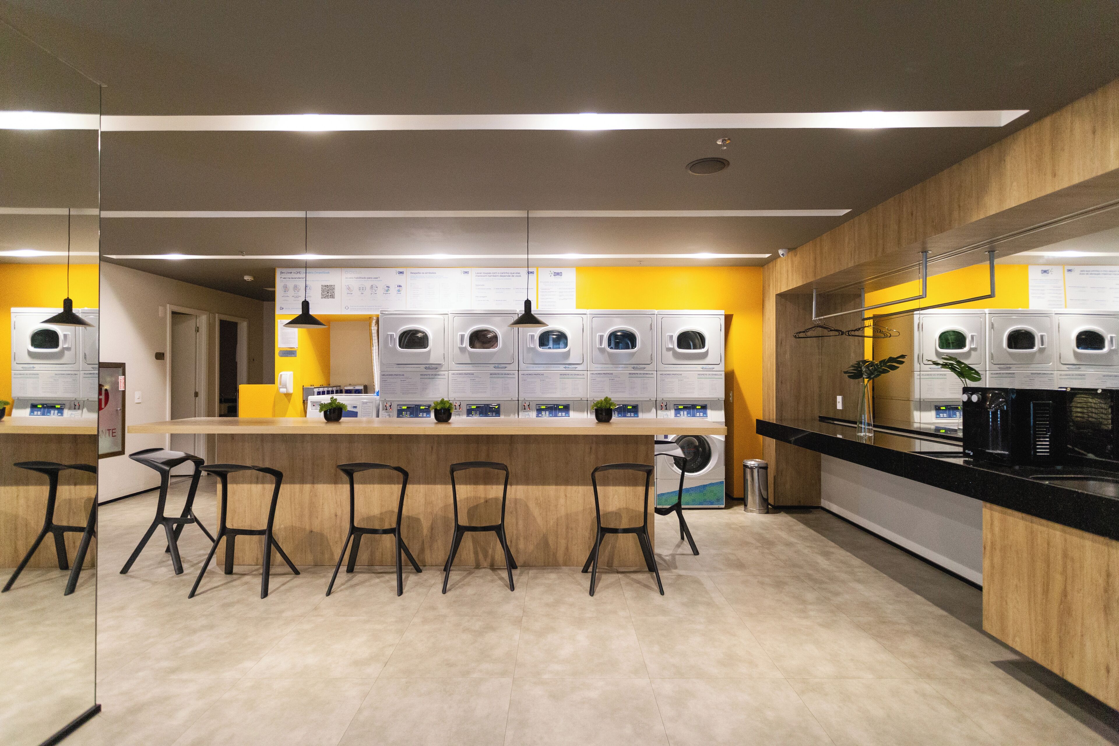A modern laundromat with multiple washing machines, a bar area with stools, and a counter with a coffee machine.