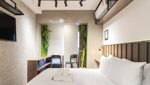 A modern hotel room with a large bed, a flat-screen TV mounted on the wall, a desk with a chair, and a small cactus plant.