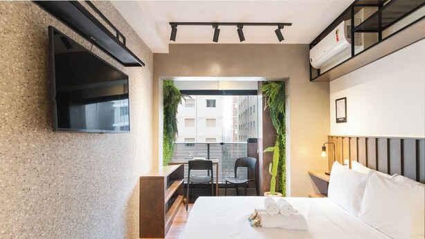 A modern hotel room with a flat-screen TV, a bed with white linens, a desk with two chairs, and a view of the city through a window.