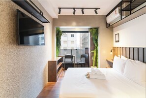A modern hotel room with a flat-screen TV, a bed with white linens, a desk with two chairs, and a view of the city through a window. - Haus Mitre Vila Mariana (São Paulo)