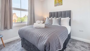 A bedroom with a large bed, a window with curtains, a bedside table, and a painting on the wall.