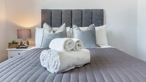 A neatly made bed with a grey quilt, white pillows, and rolled white towels.