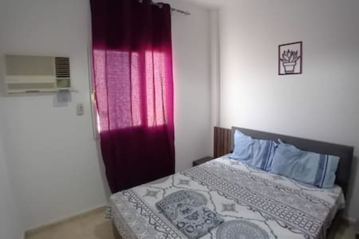 A bedroom with a bed, a window with curtains, a wall-mounted air conditioner, and a small framed picture.