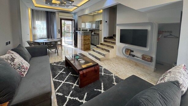 A modern living room with a sofa, coffee table, and TV. There is a kitchen area with a dining table and chairs. - 129 · 2BR Townhouse, 2 Bath, TV, Backyard (Sheikh Zayed City)