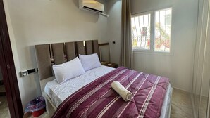 A bedroom with a bed, a ceiling fan, an air conditioning unit, a window with curtains, and a red trash bin. - 129 · 2BR Townhouse, 2 Bath, TV, Backyard (Sheikh Zayed City)