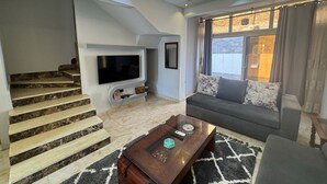 A modern living room with a staircase, a flat-screen TV, a sofa, and a coffee table. - 129 · 2BR Townhouse, 2 Bath, TV, Backyard (Sheikh Zayed City)
