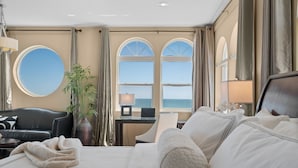 Hotelkamer met een groot bed, een bureau, een stoel, een ronde spiegel en uitzicht op de zee door boogvensters.