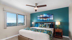 A bedroom with a bed, two bedside tables, a ceiling fan, and a large window. - Kailua Village#705 Top Floor, Gorgeous Remodeled 2 bedroom, in Heart of Town (Kailua Kona)