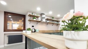 A modern kitchen with wooden countertops, a built-in stove, and a window with blinds. There are potted plants on the windowsill and a vase with flowers on the counter.