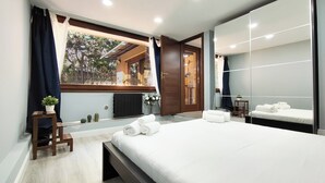 A modern bedroom with a large bed, a window with curtains, a small table with a plant, and a mirror on the wall.